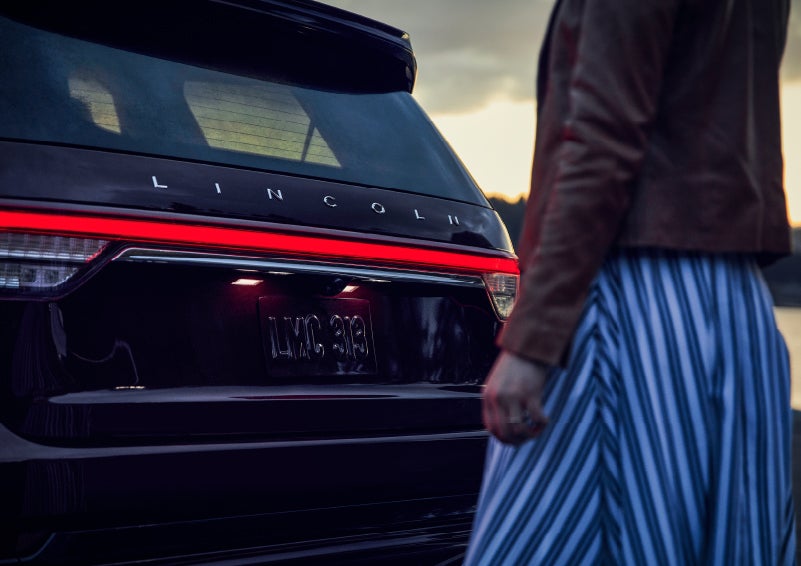 A person is shown near the rear of a 2024 Lincoln Aviator® SUV as the Lincoln Embrace illuminates the rear lights | Doral Lincoln in Doral FL