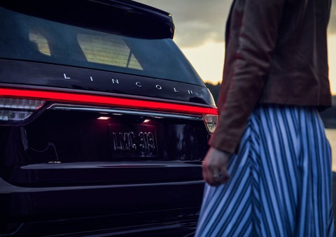 A person is shown near the rear of a 2024 Lincoln Aviator® SUV as the Lincoln Embrace illuminates the rear lights | Doral Lincoln in Doral FL