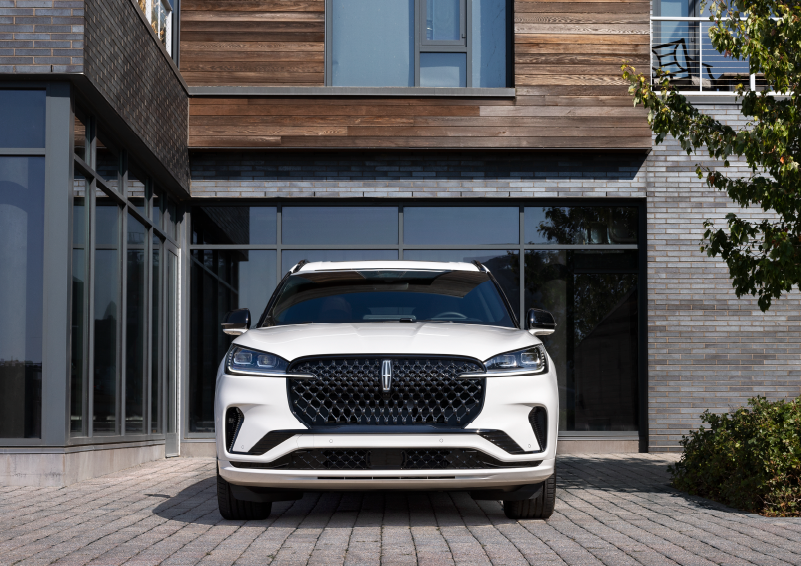 A white 2025 Lincoln Aviator® SUV is shown parked outside a modern building with gray brick and wood accents. | Doral Lincoln in Doral FL