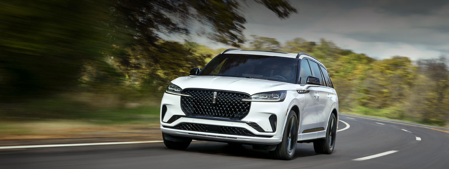 A white 2025 Lincoln Aviator® SUV is being driven down a road lined with trees. | Doral Lincoln in Doral FL